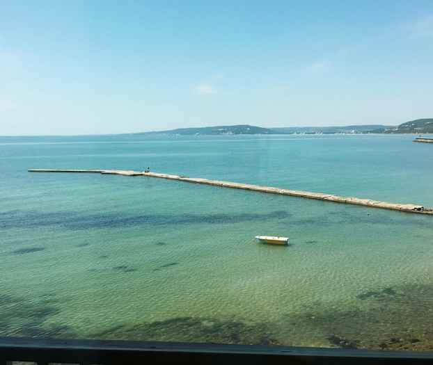 En bro strækker sig ud i det klare blå vand. En ensom båd ligger fortøjet på overfladen. (AI-genereret tekst)