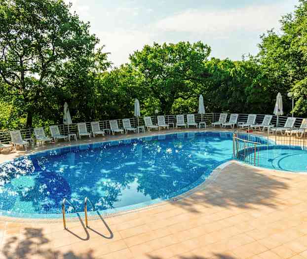 Billede af en udendørs swimmingpool omgivet af liggestole og parasoller, med grønne træer i baggrunden. (AI-genereret tekst)