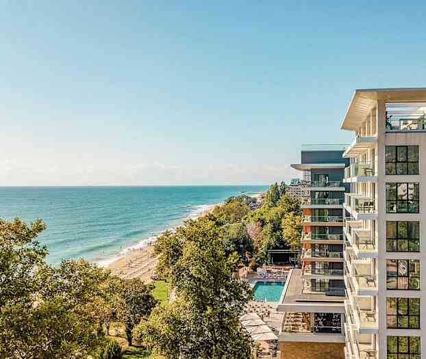 En udsigt fra højden over kysten med et hotel til højre og stranden omgivet af træer. (AI-genereret tekst)