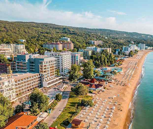 Billede af en kystby med sandstrand, parasoller og bygninger ved frodige bakker. (AI-genereret tekst)
