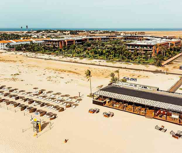 Luftfoto af en sandstrand med parasoller og liggestole samt en strandrestaurant med borde og stole omgivet af resortsbygninger og grønne planter. (AI-genereret tekst)