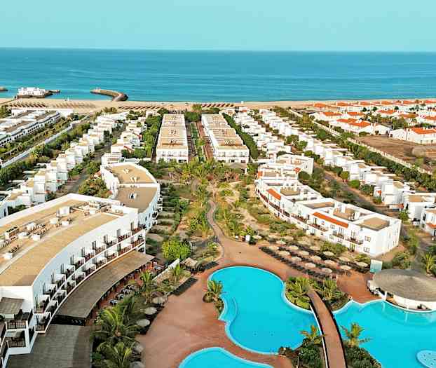 Luftfoto af et hotelområde med swimmingpools, bygninger og strand i baggrunden. (AI-genereret tekst)