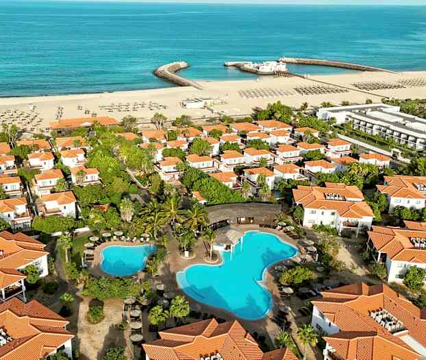 Luftfoto af et ferieområde med hvide huse med orange tage, swimmingpool og en sandstrand ved havet. (AI-genereret tekst)