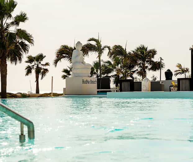 En udendørs pool med en siddende statue og omkringliggende pergolaer og palmer. (AI-genereret tekst)