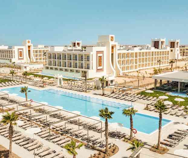 En udsigt over et feriested med swimmingpools, liggestole og omgivet af hvide bygninger. (AI-genereret tekst)