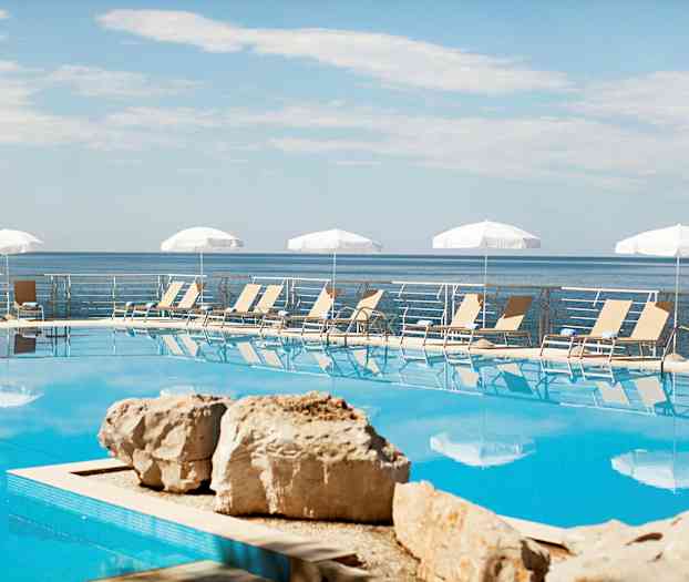 En udendørs pool med liggestole og parasoller i baggrunden, havet ses bagved poolområdet. (AI-genereret tekst)