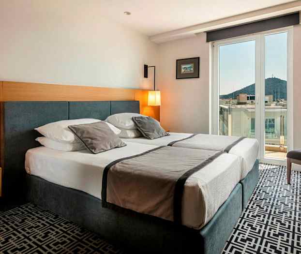 Hotelværelse med dobbeltseng, lys indretning og balkon med udsigt over by og bjerge. (AI-genereret tekst)