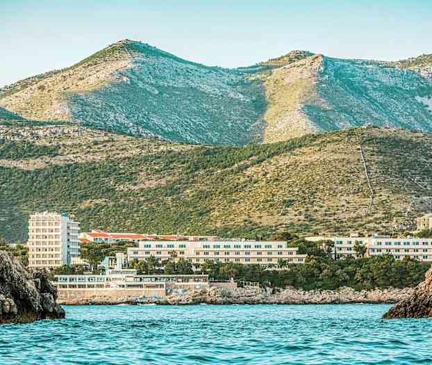 En kystudsigt med havvand i forgrunden, bygninger i midten og grønne bjerge i baggrunden. (AI-genereret tekst)