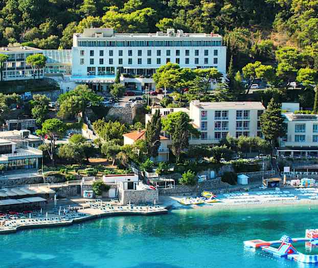 En udsigt over en hotelbygning ved en kyst med liggestole på stranden og en flydende legeplads i vandet. (AI-genereret tekst)