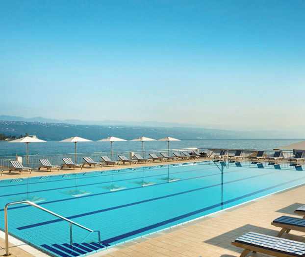 En udendørs pool med liggestole og parasoller langs kanten. I baggrunden ses havet og bjergene. (AI-genereret tekst)