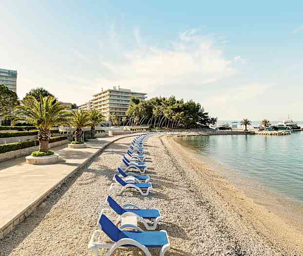 En stenstrand med solsenge og palmer, omgivet af hotelbygninger og træer. (AI-genereret tekst)