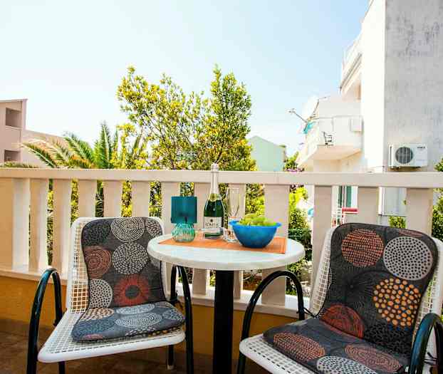 To stole med mønstrede puder står ved et lille rundt bord på en terrasse med udsigt til de omkringliggende bygninger og frodighed. (AI-genereret tekst)
