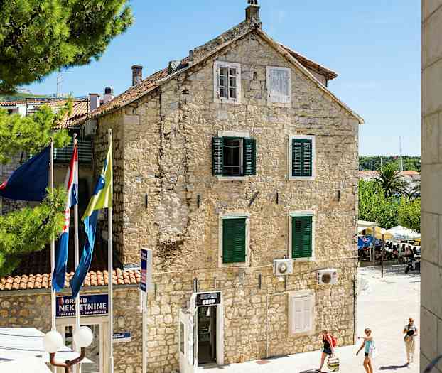 En stenbygning med grønne skodder og flag foran, omgivet af træer og mennesker, der går forbi. (AI-genereret tekst)