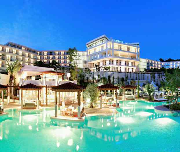 Et oplyst hotelkompleks med flere etager ved en pool, omgivet af haver og palmer, i skumringen. (AI-genereret tekst)