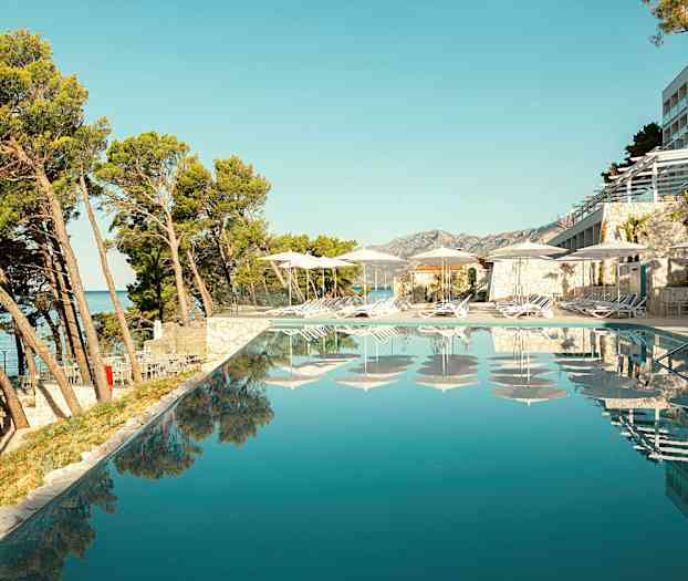 Udsigt over en infinitypool med liggestole og parasoller ved kanten, omgivet af træer og bygninger. (AI-genereret tekst)