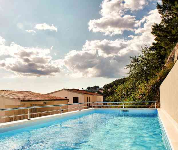 Billede af en udendørs swimmingpool med udsigt over hustage og træer under en skyet himmel. (AI-genereret tekst)