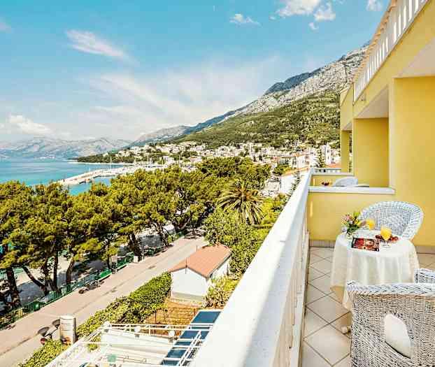 Udsigt fra en balkon på en gul bygning over en by ved havet og omkringliggende bjerge. (AI-genereret tekst)