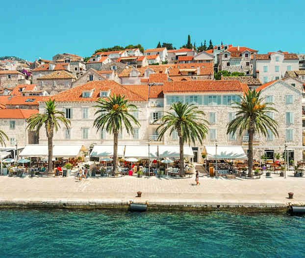 Billede af en by ved vandet med teglrøde tage og palmer langs strandpromenaden. (AI-genereret tekst)