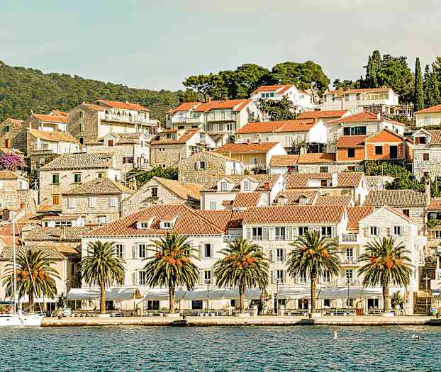 Billedet viser en pittoresk by ved havet med røde teglstenstage, stenhuse og palmer langs kajen. (AI-genereret tekst)