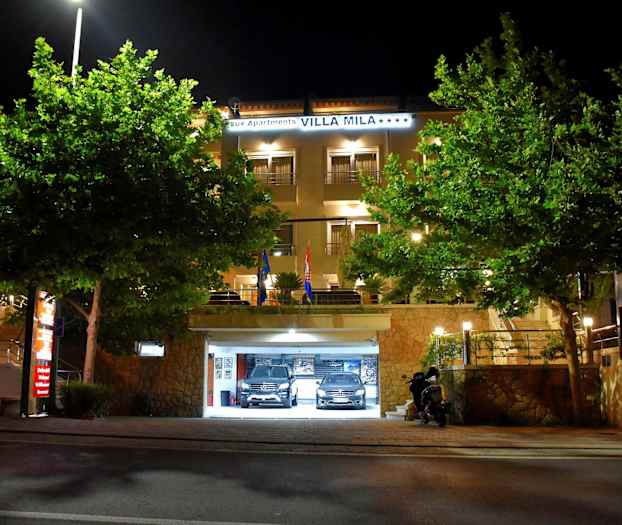 Hotelbygning med oplyst indgang og parkeringsgarage, omgivet af træer, om natten. (AI-genereret tekst)