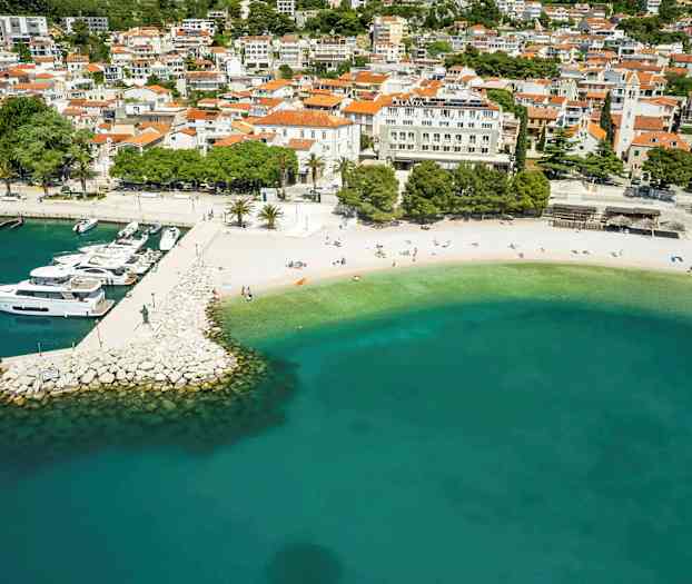Luftfoto af en strand med turkis vand og en bådebro med både, omgivet af bygninger. (AI-genereret tekst)