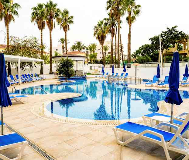 Billede af en udendørs pool omgivet af liggestole, blå parasoller og palmer. (AI-genereret tekst)