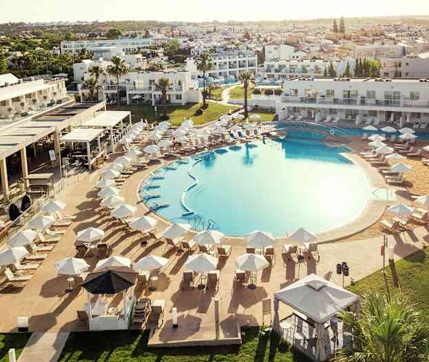 En stor udendørs pool omgivet af liggestole og parasoller, med hotelbygninger i baggrunden. (AI-genereret tekst)