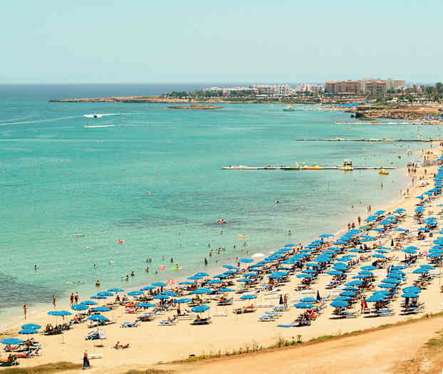 Udsigt over en strand med blå parasoller og badegæster ved det klare blå hav. (AI-genereret tekst)