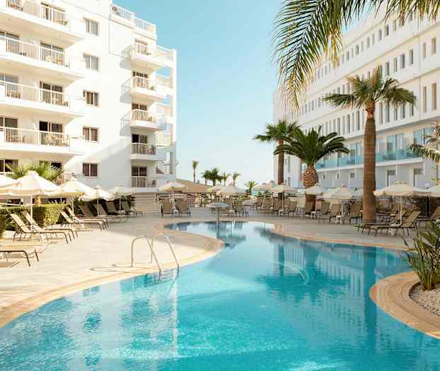 Udendørs pool med liggestole og parasoller, omgivet af hvide bygninger og palmer. (AI-genereret tekst)