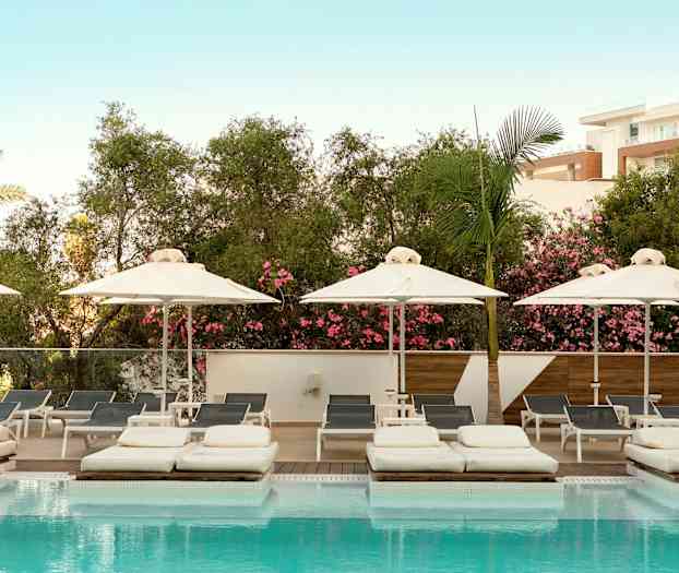 Et billede af en swimmingpool med liggestole og parasoller, omgivet af frodig vegetation og en lys blå himmel. (AI-genereret tekst)