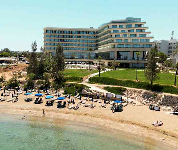 Billede af en sandstrand med opstillede parasoller og liggestole foran en moderne hotelbygning og grønne områder. (AI-genereret tekst)