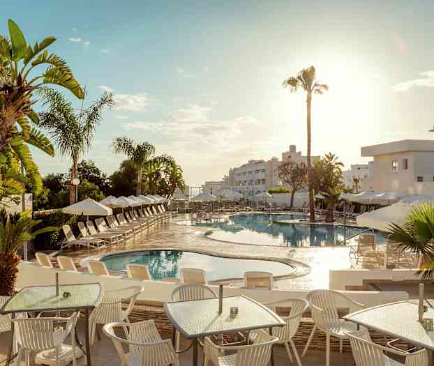 Billedet viser en udendørs swimmingpool på et hotel, omgivet af solsenge og parasoller. I forgrunden er der borde og stole under en solrig himmel. (AI-genereret tekst)