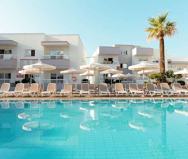 Billede af en pool med liggestole og parasoller foran et hotel. (AI-genereret tekst)