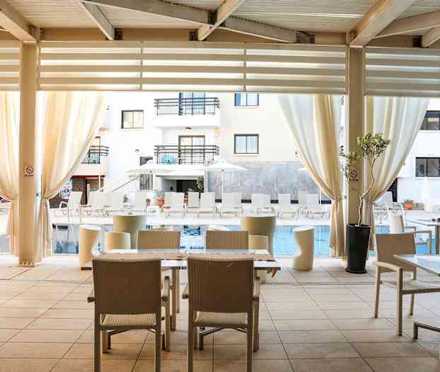 En udendørs terrasse med borde og stole under en overdækket struktur med udsigt til en pool med liggestole og parasoller. (AI-genereret tekst)