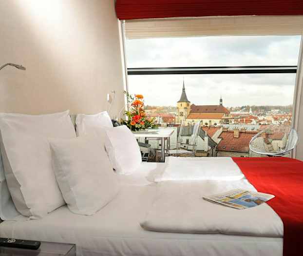Et hotelværelse med en dobbeltseng, dekoreret med rødt og hvidt sengetøj. En panoramaudsigt over en by med kirkebygninger ses gennem vinduet. (AI-genereret tekst)