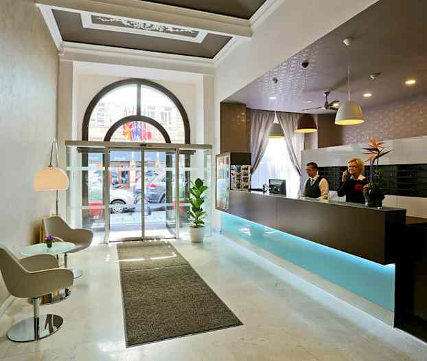 Hotellobby med reception, siddeområde og glasdøre mod ydersiden. (AI-genereret tekst)