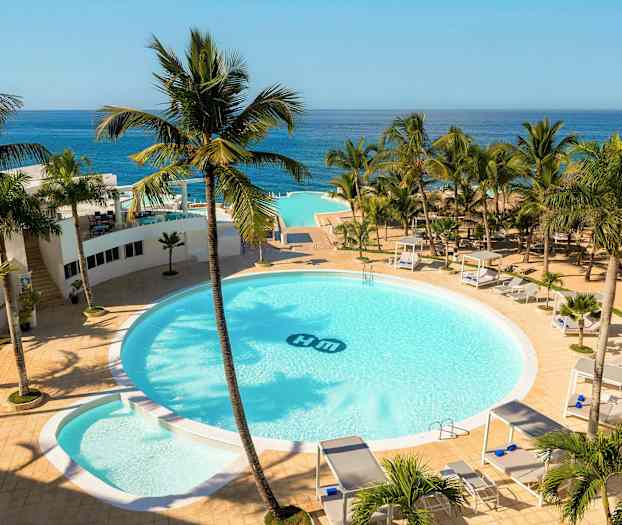 Et hotelområde med en stor rund pool omgivet af palmer, solsenge og cabanaer. Havudsigt i baggrunden. (AI-genereret tekst)