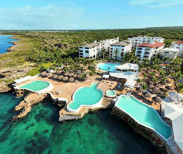 Luftfoto af en ferieanlæg med swimmingpools og baldakiner ved havet. (AI-genereret tekst)