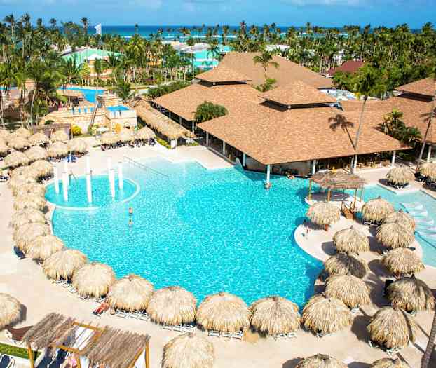 Et hotelområde med en stor pool omgivet af solsenge med palmeparaplyer og bygninger i baggrunden. (AI-genereret tekst)