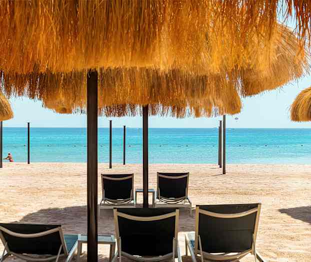 En sandstrand med liggestole og parasoller med halmtag, samt havet i baggrunden. (AI-genereret tekst)