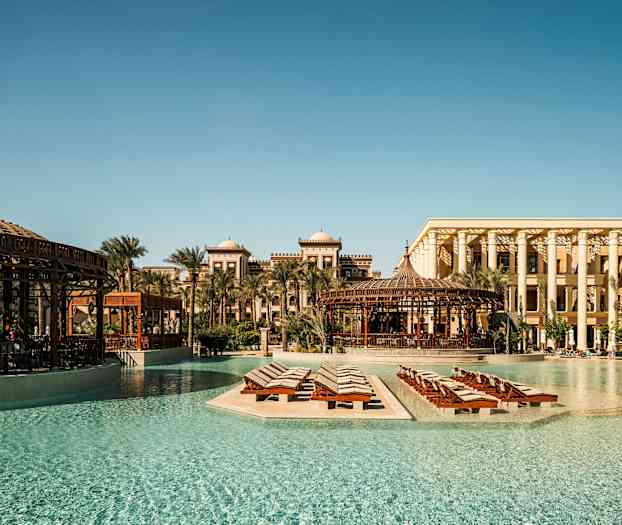 Et postkort-lignende udendørsbillede af et luksushotel med pool og liggestole omgivet af palmer og traditionel arkitektur. (AI-genereret tekst)