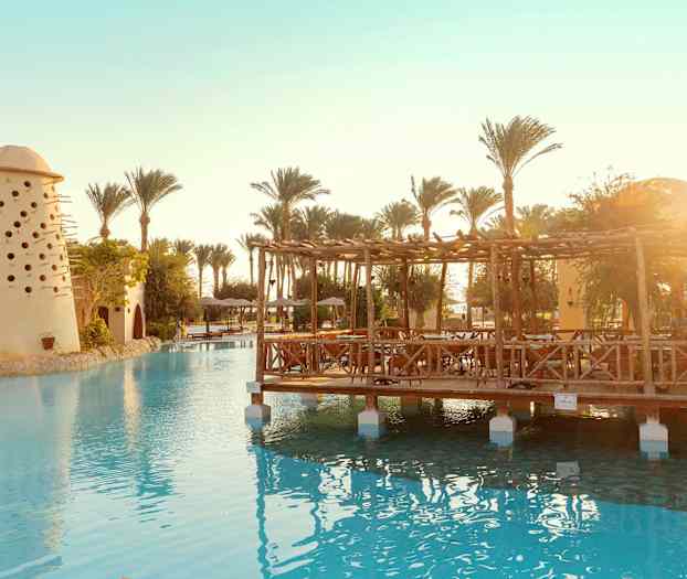 Billede af en swimmingpool med en træpavillon og en bygning med små huller, omgivet af palmer. (AI-genereret tekst)