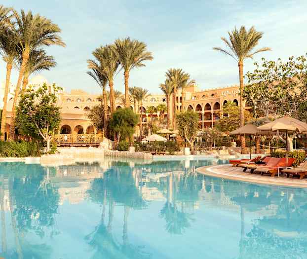 En stor pool omgivet af liggestole, parasoller, palmer og en bygning i baggrunden. (AI-genereret tekst)