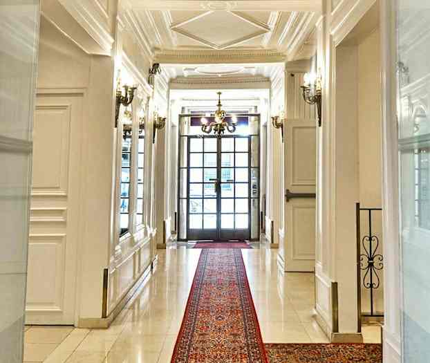 En elegant gang med hvide vægge og gulv, et rødt tæppe samt en lysekrone og glasdøre i baggrunden. (AI-genereret tekst)