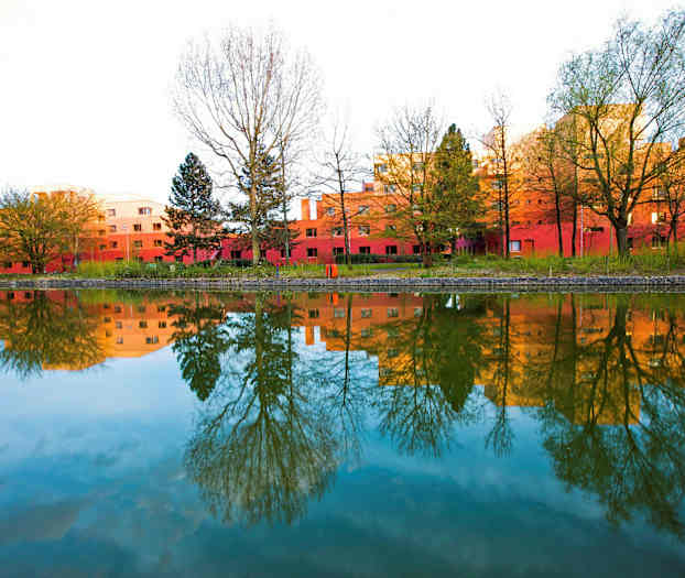 Et billede af røde bygninger omgivet af træer, der spejles i en rolig vandoverflade. (AI-genereret tekst)