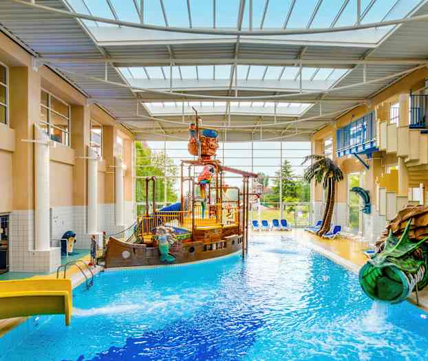 En indendørs pool med et piratskib og vandrutsjebaner, omgivet af liggestole og planter. (AI-genereret tekst)