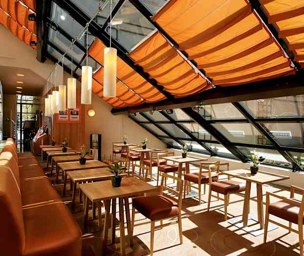 En lys og moderne spisesal med træborde og stole, orange loftsdekorationer og store vinduer. (AI-genereret tekst)