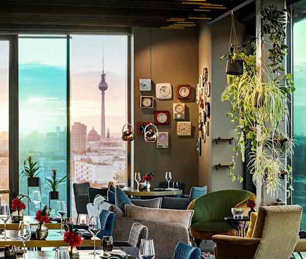 Et elegant restaurantinteriør med moderne møbler, grønne planter og udsigt over en bysilhuet med et vartegn i baggrunden. (AI-genereret tekst)