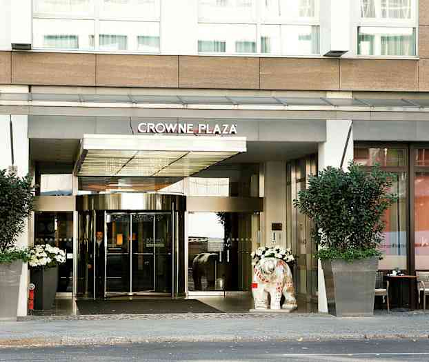 Indgangen til hotellet Crowne Plaza med glasdøre og store blomsterkrukker. (AI-genereret tekst)