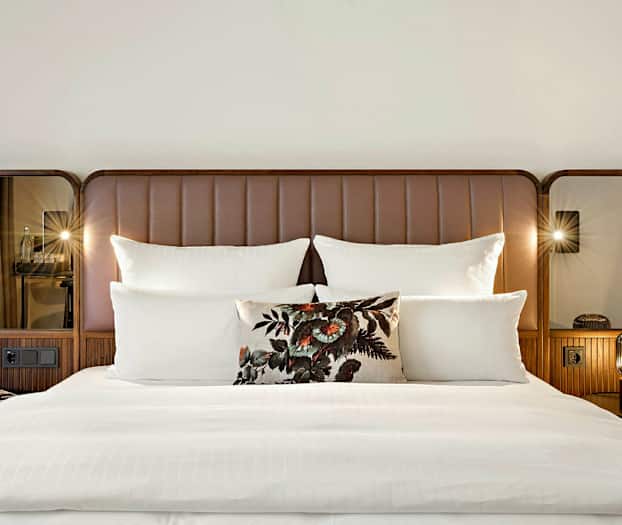 En dobbeltseng med hvidt sengetøj, flere puder og en dekorativ pude med blomsterdesign. Sengegavlen er brun med spejle på siderne. (AI-genereret tekst)